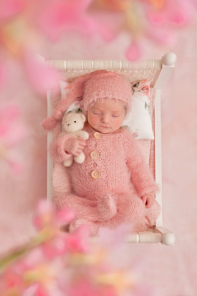 Newborn baby girl in a pink knitted romper and hat cuddling a white knitted teddy sleeping on a newborn bed and floral pillow with pink flowers and cherry blossom above