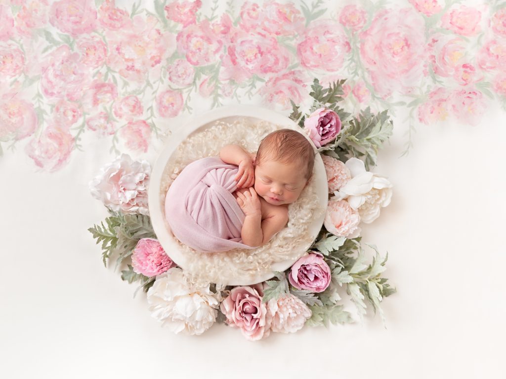 Newborn baby girl in a white bowl with delicate pink and white peony flowers on a bespoke hand painted backdrop by Horsham Photographer