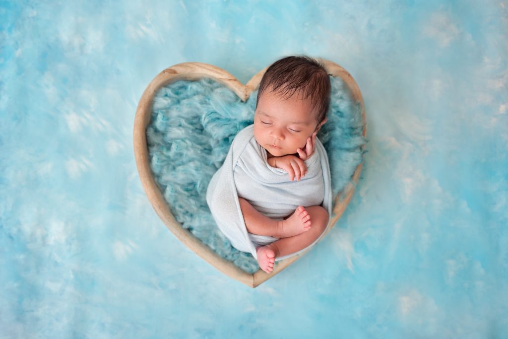 Newborn baby boy in a heart shaped wooden bowl on a hand painted bespoke blue backdrop by Horsham Photographer