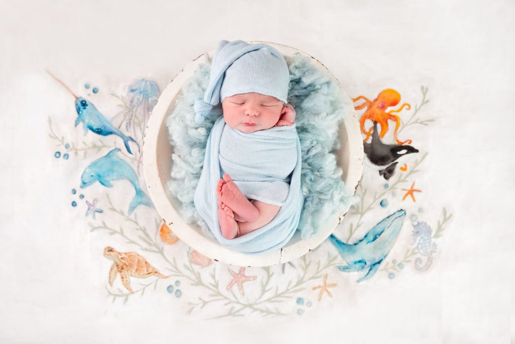 Newborn baby boy in a blue wrap and hat curled up in a bowl on a hand painted saline theme backdrop by Cate Dwyer Photography based in Horsham